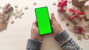 Elinde dikey yeşil ekranlı akıllı telefon tutan ve Noel arkaplanına hediyelerle dokunan bir adam. Chroma Key 'li cep telefonu. Ellerini kapat..