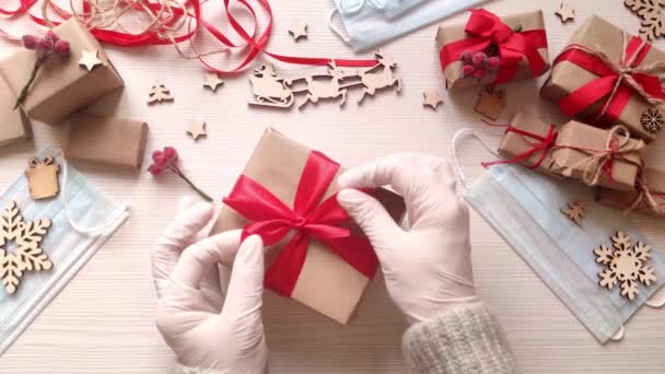 医療用手袋の女性の手は 赤いリボンでクラフト紙に存在クリスマス休暇を包みます コロナウイルスの流行の間にクリスマスボックスを梱包します ギフトや医療用マスク付きの木製テーブル — ストック動画