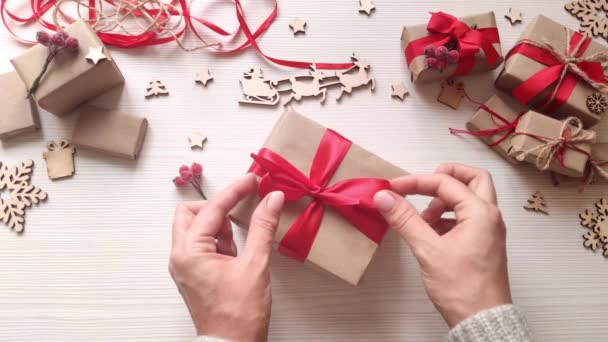 女人的手把圣诞礼物包在用红丝带和浆果做成的手工纸上 在手工制作的圣诞礼品盒上鞠躬 木制餐桌 有节日礼物 木片和星星 顶部视图 — 图库视频影像