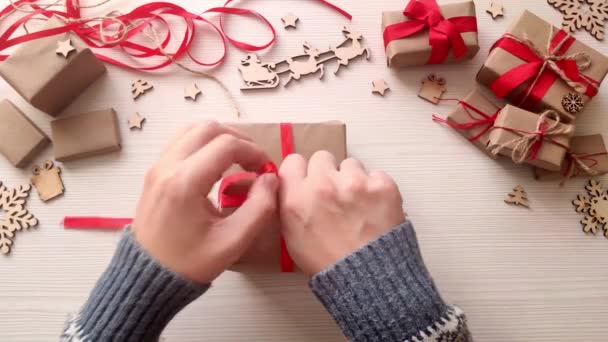 男人的手把圣诞礼物包在用红丝带和浆果做成的工艺用纸上 在手工制作的圣诞礼品盒上鞠躬 木制餐桌 有节日礼物 木片和星星 顶部视图 — 图库视频影像