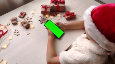 Noel Baba şapkalı şirin kız elinde dikey yeşil ekranlı akıllı telefon ve Noel arkaplanına hediyelerle dokunuyor. Tatil konsepti. Chroma Key 'li cep telefonu. Ellerini kapat..