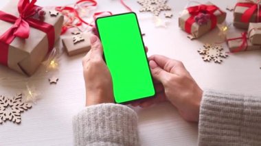 Elinde yeşil ekran olan akıllı telefonu tutan ve Noel arkaplanına hediyelerle dokunan bir kadın. Chroma Key 'li cep telefonu. Ellerini kapat..