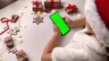 Noel Baba şapkalı şirin kız elinde dikey yeşil ekranlı akıllı telefon, ekrana dokunuyor ve hediyelerle Noel arkaplanına kayıyor. Tatil konsepti. Chroma Key 'li cep telefonu. Ellerini kapat..