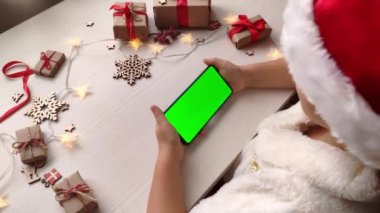 Noel Baba şapkalı şirin kız elinde yatay yeşil ekranlı akıllı telefon ve Noel arkaplanına hediyelerle dokunuyor. Tatil konsepti. Chroma Key 'li cep telefonu. Ellerini kapat..