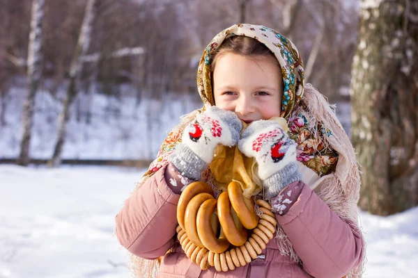 冬の背景にパンケーキやベーグルを持つ伝統的なロシアのスカーフでかわいい女の子 民俗服の子供のクローズアップ肖像画 マレニツァ祭 シュロヴェチドのための伝統的なロシア料理 — ストック写真