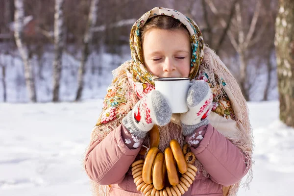 冬の背景に熱いお茶とバゲルのマグカップと伝統的なロシアのスカーフでかわいい女の子 民俗服の子供のクローズアップ肖像画 マレニツァ祭 — ストック写真