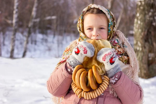 冬の背景にパンケーキやベーグルを持つ伝統的なロシアのスカーフでかわいい女の子 民俗服の子供のクローズアップ肖像画 マレニツァ祭 シュロヴェチドのための伝統的なロシア料理 — ストック写真