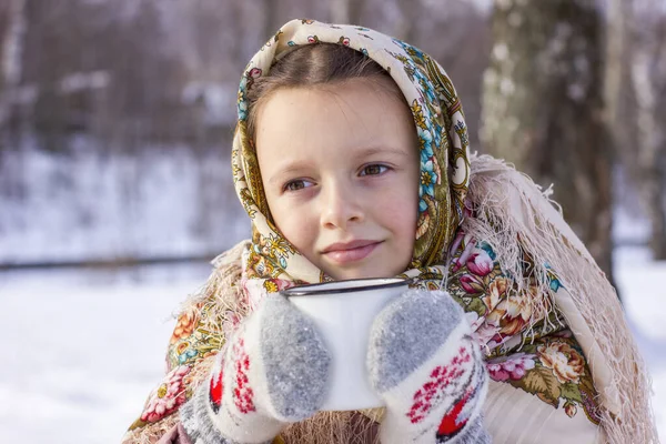 冬の背景に熱いお茶のマグカップと伝統的なロシアのスカーフでかわいい女の子 民俗衣装に身を包んだ子供の肖像 — ストック写真