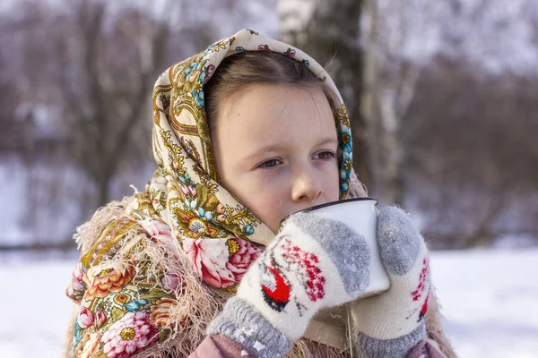 冬の背景に熱いお茶のマグカップと伝統的なロシアのスカーフでかわいい女の子 民俗衣装に身を包んだ子供の肖像 — ストック写真