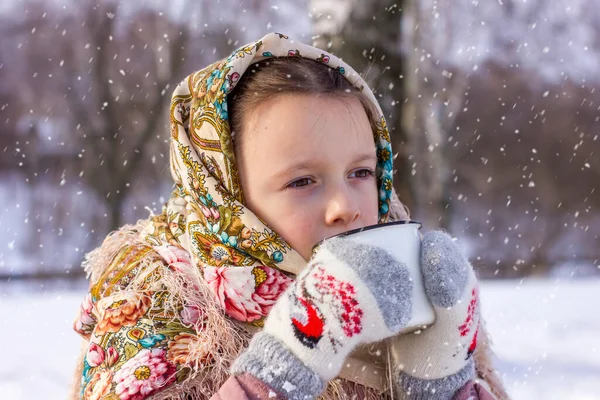 冬の背景に熱いお茶のマグカップと伝統的なロシアのスカーフでかわいい女の子 民俗衣装に身を包んだ子供の肖像 — ストック写真