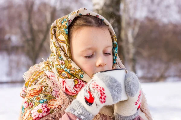 冬の背景に熱いお茶のマグカップと伝統的なロシアのスカーフでかわいい女の子 民俗衣装に身を包んだ子供の肖像 — ストック写真