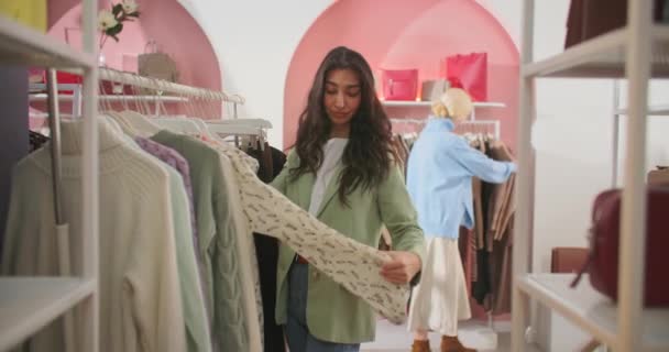 Jonge aantrekkelijke Brunette vrouw onderzoekt zorgvuldig kleding op een rek of Hanger in een Shopping Center Store. Portret van een mooi meisje raakt en horloges verkoop — Stockvideo