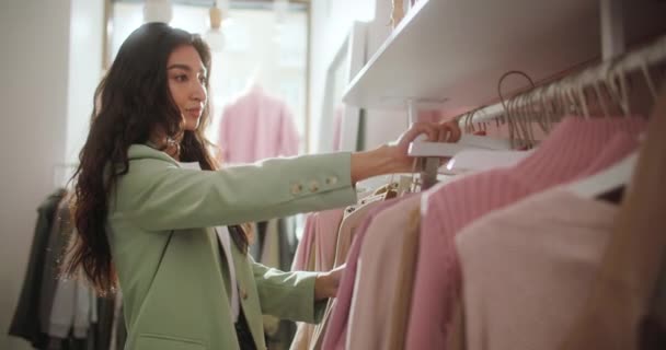 Ung attraktiv brunett kvinna undersöker omsorgsfullt kläder på en rack eller Hanger i ett köpcentrum butik. Porträtt av en söt flicka berör och klockor försäljning — Stockvideo