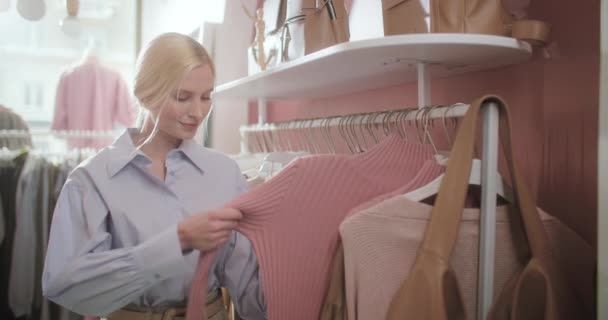 Hezké mladé blondýny žena výběru oblečení v obchodě s oblečením. Žena Zákazník v ležérní oblečení Výběr oblečení z regálu, police v obchodním domě. — Stock video