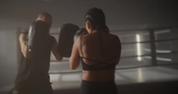 O tânără sportivă și un bărbat caucazian se antrenează în flăcări pe inelul de box. Sparring of Young Sportswoman and Personal Trainer in Backlight. Combat Sport, Stil de viață . — Videoclip de stoc