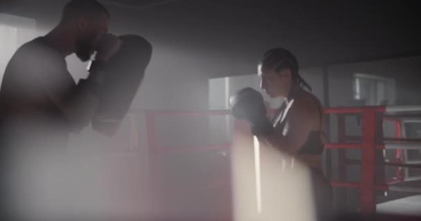 Молодая спортсменка в боксерских перчатках и спортивной одежде боксирует со своим умелым тренером. Они избегают ударов, тренируясь на боксёрском ринге. — стоковое видео