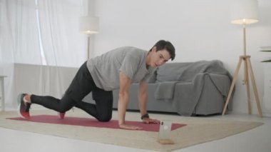 Gri tişörtlü yakışıklı genç adam ve siyah şortlu adam internette spor yapıyor. Güçlü Atletik Erkek Geniş ve Güneşli Oturma Odasında Çapraz Çatırtı Egzersizleri Yapıyor.