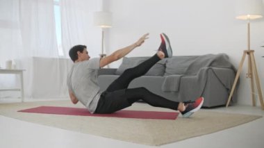 Gri tişörtlü yakışıklı genç adam ve siyah şortlu adam internette spor yapıyor. Güçlü Atletik Erkek Geniş ve Güneşli Oturma Odasında Çapraz Çatırtı Egzersizleri Yapıyor.