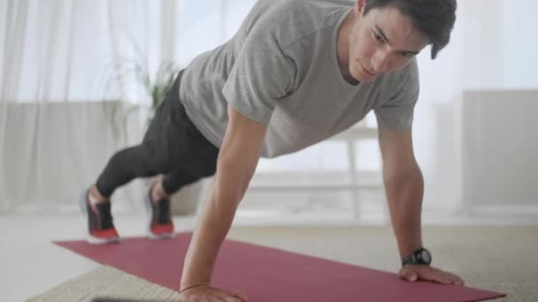 Snygg ung man i en grå T-shirt och svarta shorts gör sport hemma på nätet. Attraktiva manliga gör Plank Motion på Hhome under karantän. — Stockvideo