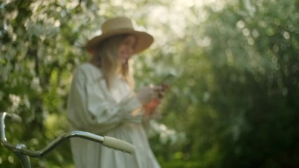 Πορτρέτο της ξανθιάς γυναίκας στο καπέλο Απολαύστε την επιτυχία στο κινητό τηλέφωνο του White Apple Blossoms. Κήπος με ανθισμένα δέντρα. Η έννοια της ομορφιάς, της υγείας, της φρεσκάδας και της φυσικότητας. — Αρχείο Βίντεο