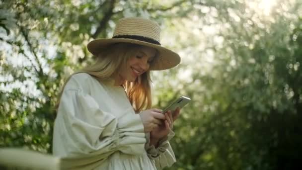 Portrait of Blonde Woman in Hat Enjoy Takes Pictures at Smartphone Device of White Apple Blossoms Сад з кульгавими деревами. Концепція краси, здоров'я, свободи і природності. Зачиніть. — стокове відео