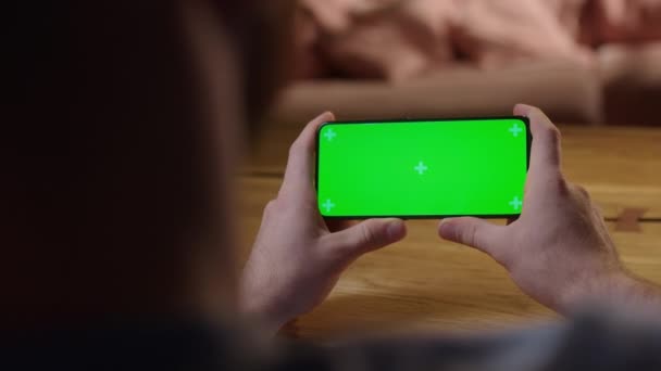 Cámara de mano: Vista trasera del joven en casa sentado en un escritorio de madera con un teléfono inteligente de pantalla falsa verde. Boy está viendo contenido sin tocar la pantalla del gadget. — Vídeos de Stock