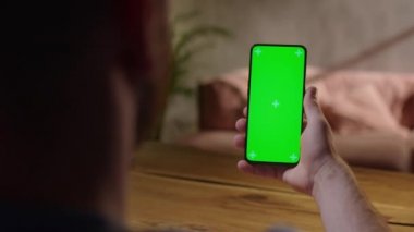 El kamerası: Young Man in Home Sitting on a Wood Desk kullanırken Green Mock-up Screen Smartphone 'u kullanıyor. Oğlan Dokunmadan İçerik Ekranı İzliyor.