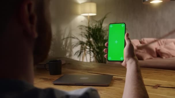 Φορητή κάμερα: Πίσω όψη του νεαρού άνδρα στο σπίτι που κάθεται σε ένα ξύλινο γραφείο χρησιμοποιώντας με πράσινο mock-up οθόνη Smartphone. Αγόρι παρακολουθεί περιεχόμενο χωρίς να αγγίζει Gadget οθόνη. — Αρχείο Βίντεο