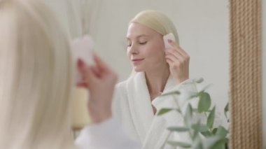 Parlak Banyo 'da yüz masajı yapan güzel bir kadın. Ağır çekim. Çekici Sarışın Kadın Portresi Guasha 'lı Bornoz Giyen Yüzüne Masaj Yapan ve Kameraya Bakan