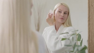 Parlak Banyo 'da yüz masajı yapan güzel bir kadın. Ağır çekim. Çekici Sarışın Kadın Portresi Guasha 'lı Bornoz Giyen Yüzüne Masaj Yapan ve Kameraya Bakan