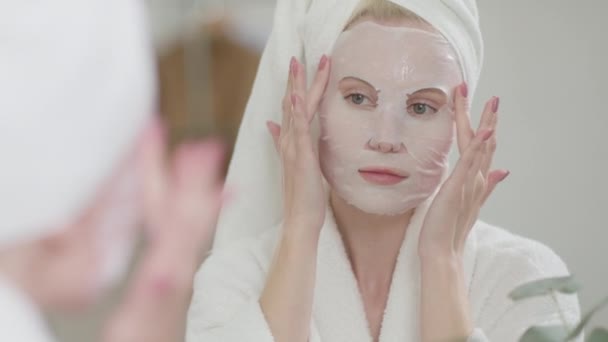 Jolie dame enveloppée dans une serviette de bain bénéficiant des procédures du matin à la maison. Femme blonde appliquant un masque japonais cosmétique sur son visage et regardant dans le miroir regardant la caméra dans la salle de bain — Video
