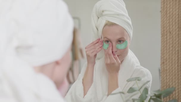 Ευτυχισμένη ελκυστική γυναίκα στο μπάνιο πετσέτα με καλλυντικά μπαλώματα κάτω από τα μάτια Kooking σε καθρέφτη και απολαμβάνοντας την πρωινή ώρα στο φωτεινό μπάνιο. — Αρχείο Βίντεο