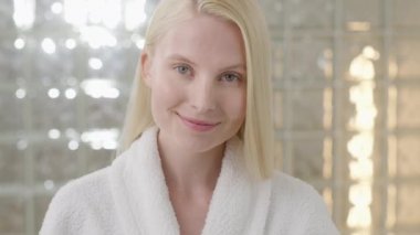 Banyo havlusu takan, uzun sarı saçlı beyaz bir kadın, parlak bir banyoda kameraya bakıyor. Cilt bakımı spa prosedürlerinin reklamını yapmak. Yakın plan portre
