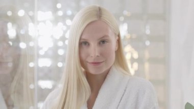 Banyo havlusu takan, uzun sarı saçlı beyaz bir kadın, parlak bir banyoda kameraya bakıyor. Cilt bakımı spa prosedürlerinin reklamını yapmak. Yakın plan portre