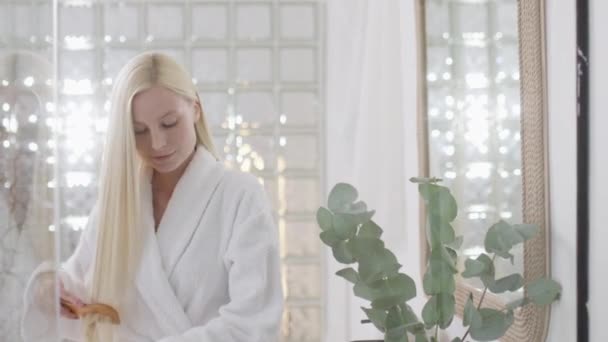 Jonge blanke vrouw met lang blond haar dragen van een badhanddoek Kijken naar de camera en kammen gezonde rechte haren in een heldere badkamer. Langzame beweging — Stockvideo