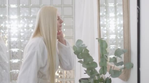 Боковой вид молодой белокурой женщины с длинными светлыми волосами, носящей банное полотенце, смотрящей в зеркало и расчесывающей здоровые прямые волосы в светлой ванной комнате. Slow Motion — стоковое видео