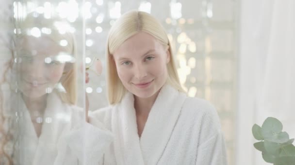 Pretty Female doing Facial Massage in Bright koupelna. Pomalý pohyb. Portrét atraktivní mladé blond ženy v županu s masážním válečkem masáže její obličej a při pohledu na kameru — Stock video