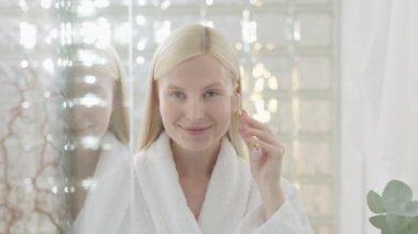 Parlak Banyo 'da yüz masajı yapan güzel bir kadın. Ağır çekim. Çekici Sarışın Kadın Portresi Roller Masajlı Bornoz Giyen Yüzüne Masaj Yapan ve Kameraya Bakan