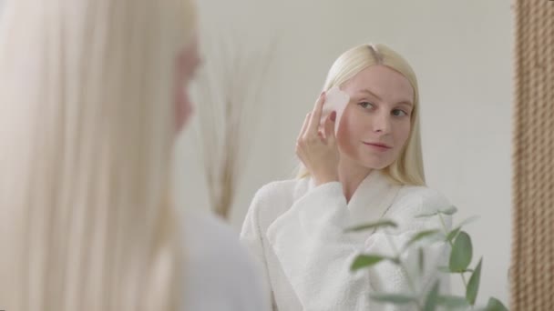 Mooie vrouw doet gezichtsmassage in lichte badkamer. Slow Motion. Portret van aantrekkelijke jonge Blond vrouw dragen badjas met een Guasha massages haar gezicht en kijken naar camera — Stockvideo