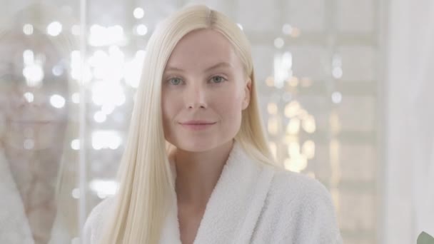 Junge kaukasische Frau mit langen blonden Haaren, die ein Badetuch trägt und in einem hellen Badezimmer in die Kamera blickt. Werbung für das Konzept der Hautpflege-Wellness-Verfahren. Nahaufnahme — Stockvideo