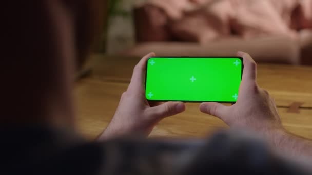 Cámara de mano: Hombre joven en el hogar que sostiene la clave de croma Pantalla verde Smartphone Ver contenido sin tocar o deslizar. Navegar por Internet, Ver contenido, Vídeos — Vídeo de stock