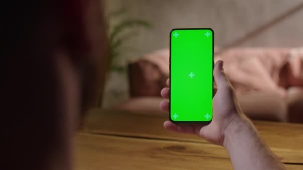 Cámara de mano: Vista trasera del joven en casa sentado en un escritorio de madera con un teléfono inteligente de pantalla falsa verde. Boy está viendo contenido sin tocar la pantalla del gadget. — Vídeo de stock