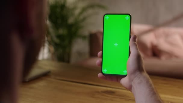 Cámara de mano: Vista trasera del joven en casa sentado en un escritorio de madera con un teléfono inteligente de pantalla falsa verde. Boy está viendo contenido sin tocar la pantalla del gadget. — Vídeos de Stock