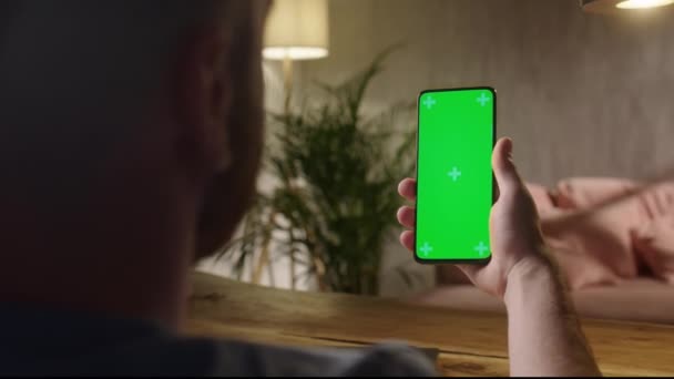 Cámara de mano: Vista trasera del hombre en la habitación moderna sentado usando el teléfono con Green Mock-up Screen Chroma Key Con Track Points Navegación por Internet Ver contenido Videos Blogs. Desliza hacia arriba — Vídeo de stock