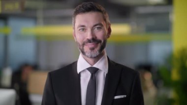 Çalışanlar arka planda çalışmakla meşgulken ofiste durup kameraya gülümseyen yetişkin bir iş adamı portresi.
