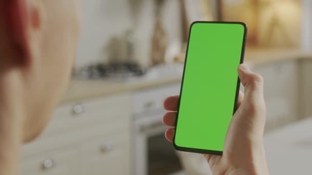 Вид ззаду молодого чоловіка вдома сидить на кухні, використовуючи з зеленим макет екрану смартфона. Хлопчик розміщує контент, не торкаючись екрана гаджетів. Модульна технологія та інформаційна концепція . — стокове відео