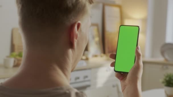 Вид ззаду молодого чоловіка вдома сидить на кухні, використовуючи з зеленим макет екрану смартфона. Хлопчик розміщує контент, не торкаючись екрана гаджетів. Модульна технологія та інформаційна концепція . — стокове відео