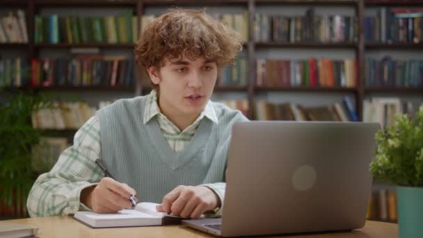 自宅や図書館でのオンラインレッスンでの若い学生の男の研究電話会議ビデオチャットを行う,ノートを作る自宅のオフィスデスクで座ってノートパソコンを見て — ストック動画