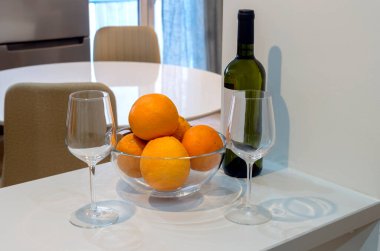 Masada iki boş bardak, bir şişe beyaz şarap ve bir kase portakal var.