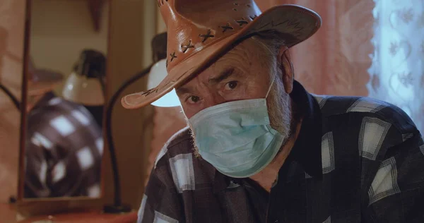 Şapkalı ve tıbbi maskeli yaşlı bir adamın portresi hüzünle kameraya bakıyor. Stok Fotoğraf